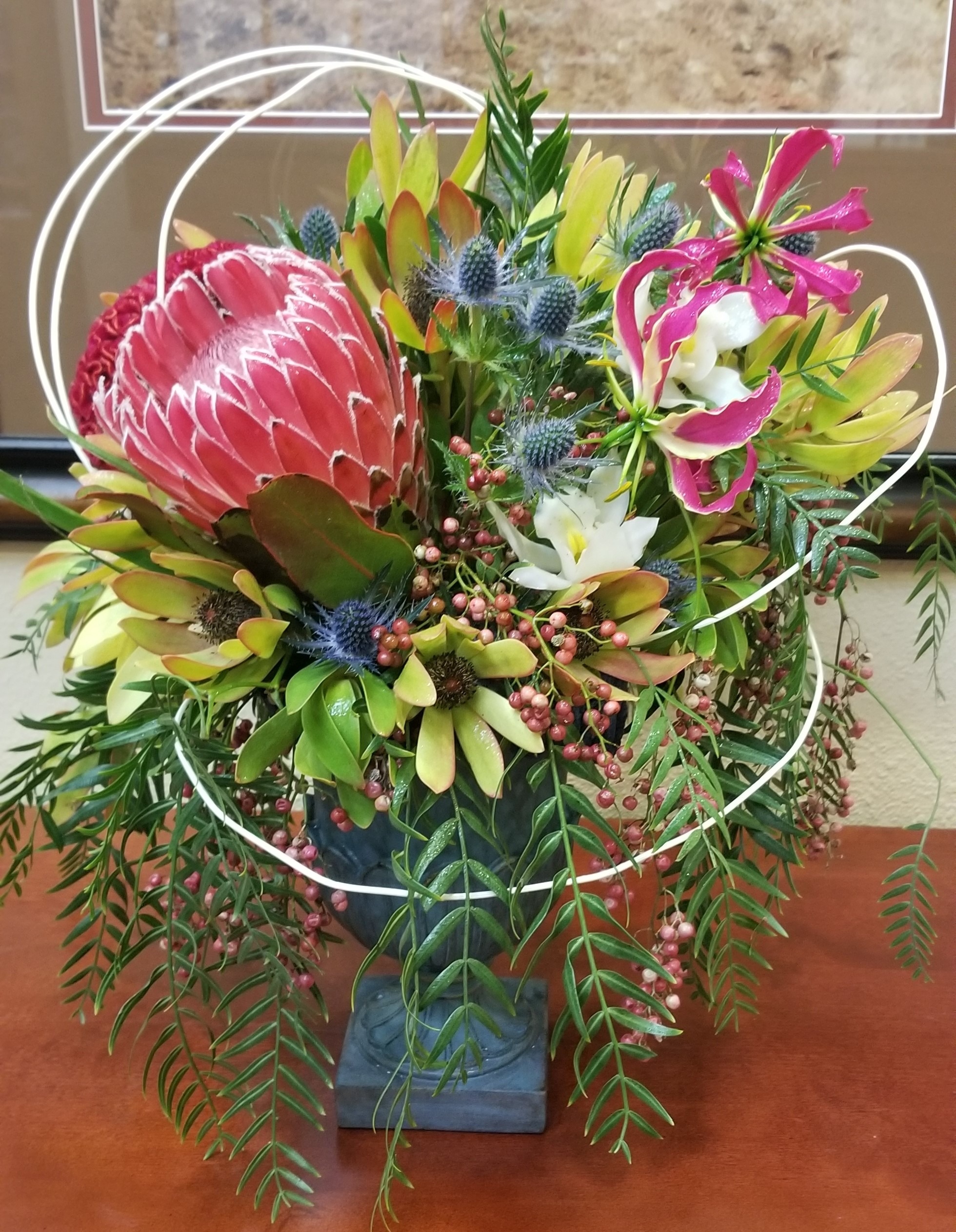 Vibrant floral arrangement featuring a large protea, various greenery, and colorful blooms, displayed in a decorative vase on a wooden surface.