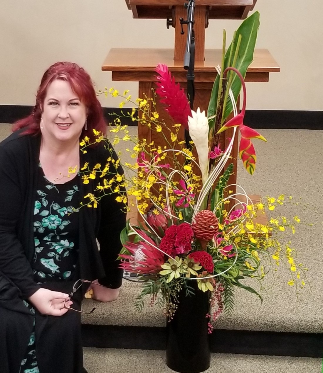 A woman with red hair poses beside a vibrant floral arrangement, featuring bright yellow and pink flowers, in a setting with a podium.