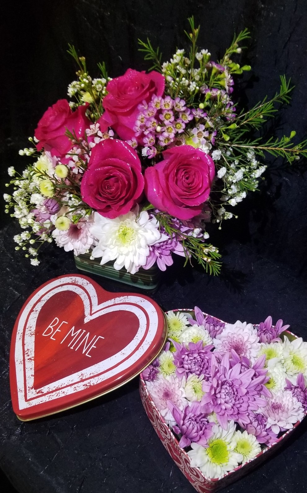 A vibrant flower arrangement featuring pink roses, white daisies, and purple chrysanthemums, alongside a heart-shaped box labeled "BE MINE," perfect for Valentine's gifts.