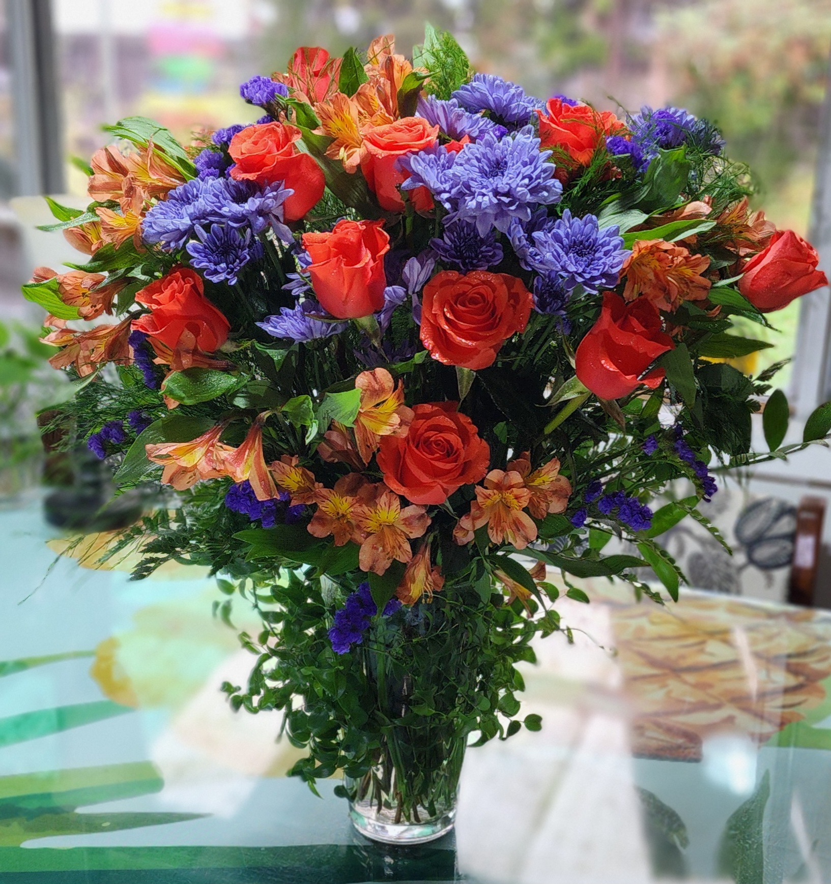 Vibrant bouquet featuring orange roses, purple asters, and yellow alstroemeria in a clear vase, set against a softly blurred background.