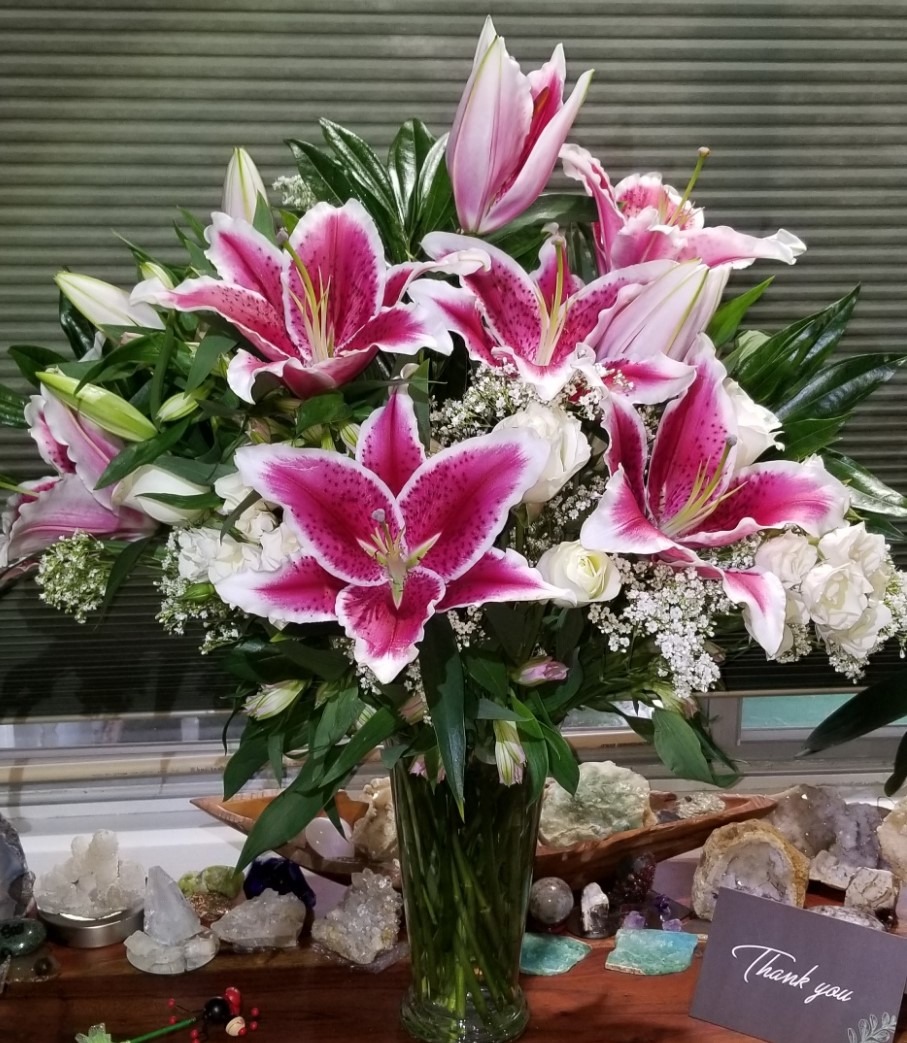 Vibrant bouquet featuring pink and white lilies, mixed with roses and greenery, displayed in a clear vase. A "Thank you" card is visible on the table.