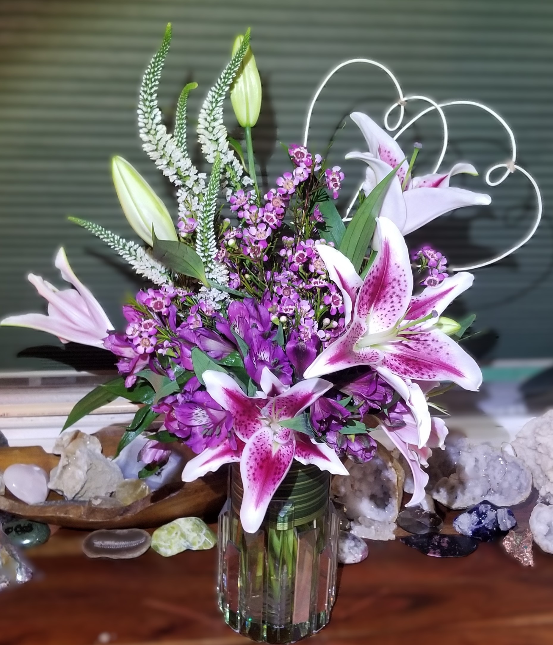 Vibrant bouquet featuring purple lilies, greenery, and delicate filler flowers, arranged in a textured glass vase, enhancing a floral centerpiece.